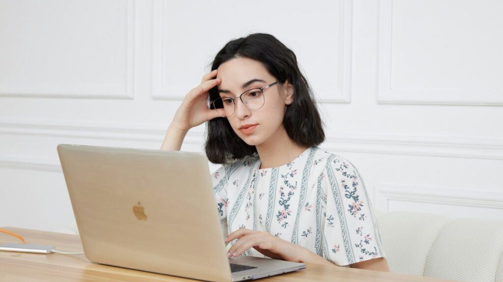 Worried young woman resting her head on her hand while looking at her laptop, feeling anxious about new technology