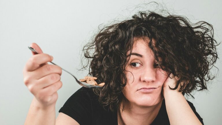 Tired woman resting her head on her hand while holding a spoonful of cereal she has no appetite to eat